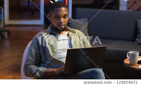Adult guy typing on laptop laying on couch at his stylish loft space, engaging in freelancing and online work. Black person multitasking and embracing the flexibility of remote job. Camera B. Adult guy typing on laptop laying on couch at his stylish loft space, engaging in freelancing and online work. Black person multitasking and embracing the flexibility of remote job. Camera B. 129854072