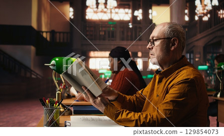 Old university student preparing his thesis in a campus library study room, reading a book for academic information. Ambition and determination to complete a college degree. 129854073