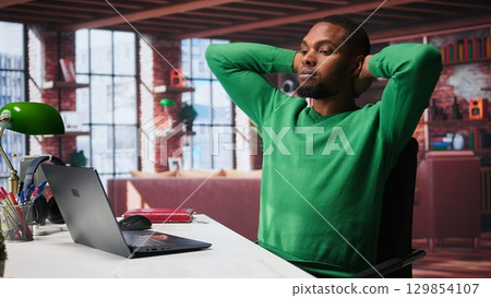 Man at home deals with back pain and stretching joints on chair, being uncomfortable due to incorrect posture. Black guy feeling overworked and experiencing muscle cramps at desk. Camera B. 129854107