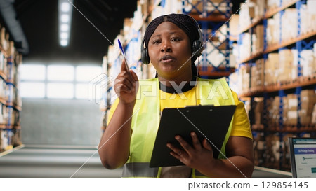 African American woman in hi vis vest handles calls on the help line, assisting people needing help with parcel tracking or other shipment information. E-commerce activity. Camera B. 129854145