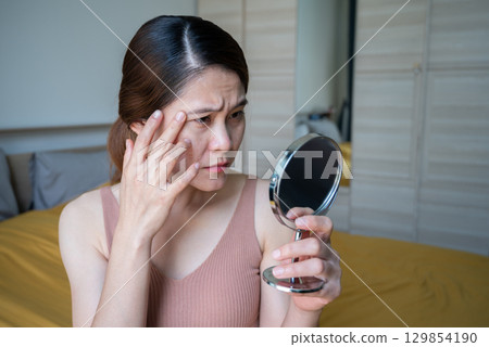 Asian woman worried about her face when she saw acne and wrinkled occur on her face by mini mirror. Allergies, irritants, genetic makeup, etc can cause skin conditions. Asian woman worried about her face when she saw acne and wrinkled occur on her face by mini mirror. Allergies, irritants, genetic makeup, etc can cause skin conditions. 129854190