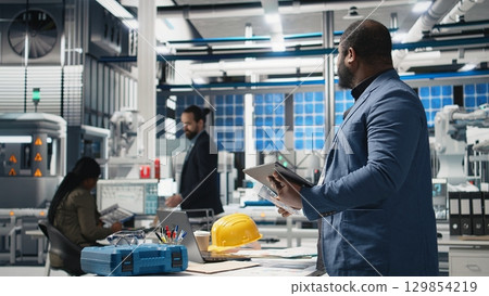 Black master engineer overseeing photovoltaics production line in a factory, ensuring a successful manufacturing process to meet corporate goals and alternative energy standards. Camera A. Black master engineer overseeing photovoltaics production line in a factory, ensuring a successful manufacturing process to meet corporate goals and alternative energy standards. Camera A. 129854219