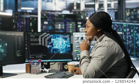Data center technician using artificial intelligence deep learning models to configure hardware. African american woman works on AI programming language in server room, optimizing system, camera B Data center technician using artificial intelligence deep learning models to configure hardware. African american woman works on AI programming language in server room, optimizing system, camera B 129854244