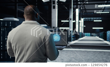 IT expert walking in server hub using laptop to enhance AI model accuracy. Data center technician using notebook to schedule routine maintenance for AI hardware components, camera A 129854276
