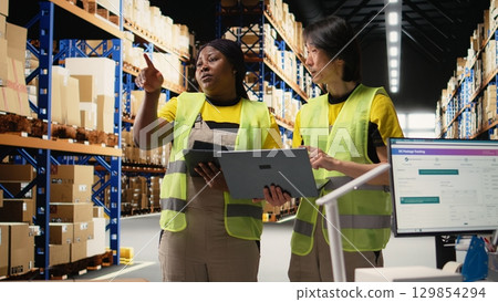 Team of diverse workers handling tracking software for parcel status on tablet, following the cargo shipment from the industrial warehouse with racks. Staff in hi vis vests. Camera A. Team of diverse workers handling tracking software for parcel status on tablet, following the cargo shipment from the industrial warehouse with racks. Staff in hi vis vests. Camera A. 129854294