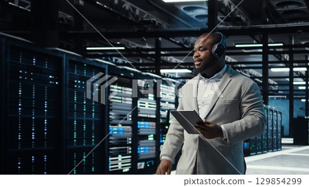 Data center admin wearing headphones using tablet to identify bottlenecks. Server farm IT expert listening music while troubleshooting bugs, implementing optimizations, camera A Data center admin wearing headphones using tablet to identify bottlenecks. Server farm IT expert listening music while troubleshooting bugs, implementing optimizations, camera A 129854299