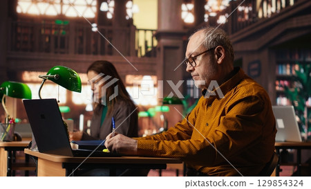 Focused senior person reading web educational papers in a university library, committed to completing his studies and achieving a meaningful degree in his later years. Old man. 129854324