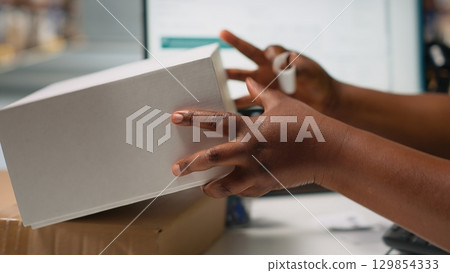 Black woman packing goods and applying shipping labels on boxes, creating awb tracking numbers on tags for easy distribution. Worker using adhesive airway bill stickers. Camera B. 129854333