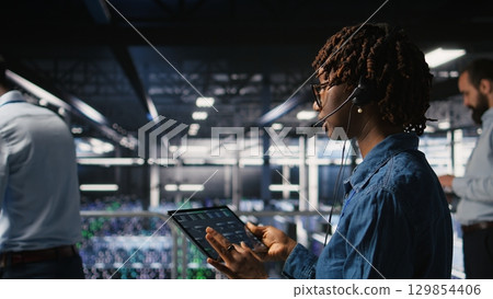 Server hub IT expert monitors systems, doing tech support, guiding users through troubleshooting steps. African american technician providing remote diagnostics using headset mic, Server hub IT expert monitors systems, doing tech support, guiding users through troubleshooting steps. African american technician providing remote diagnostics using headset mic, 129854406