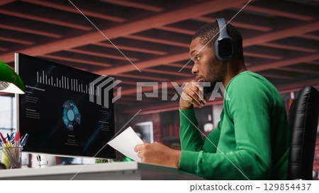 Teleworker in home office watching business conference on PC, looking over statistics charts. African american remote employee in apartment looking at presentation graphs on computer. Camera A. 129854437