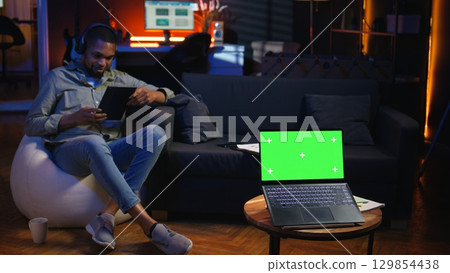 Young male person enjoys leisure time next to chroma key on bean bag, uses his smartphone to watch a podcast with headset. Black guy relaxing remotely in the living room. Camera A. Young male person enjoys leisure time next to chroma key on bean bag, uses his smartphone to watch a podcast with headset. Black guy relaxing remotely in the living room. Camera A. 129854438