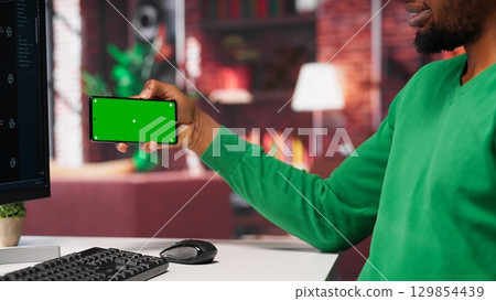 African american man engaging with AI chatbot next to chroma key, using the virtual assistant with prompts for data driven algorithms to enhance productivity at home office. Camera A. African american man engaging with AI chatbot next to chroma key, using the virtual assistant with prompts for data driven algorithms to enhance productivity at home office. Camera A. 129854439