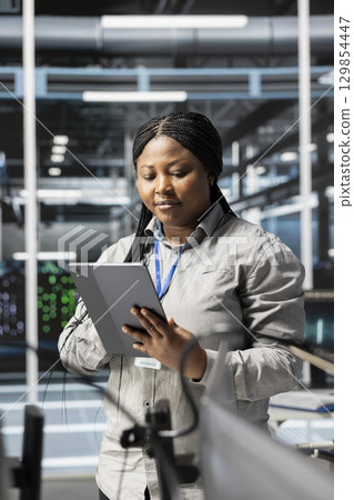 Woman in data center doing software updates, verifying deep learning parameters. African american server hub IT expert using PC to inspect configurations, ensuring optimal application performance Woman in data center doing software updates, verifying deep learning parameters. African american server hub IT expert using PC to inspect configurations, ensuring optimal application performance 129854447