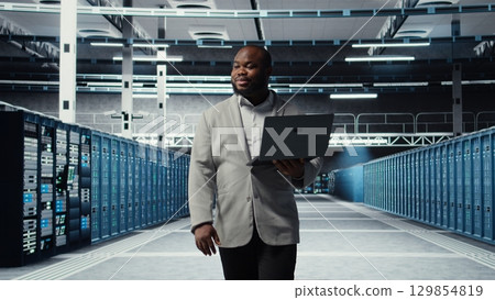 Smiling server room engineer performing maintenance on server racks using software on laptop. Happy data center IT professional upgrading blade server components using notebook, camera A Smiling server room engineer performing maintenance on server racks using software on laptop. Happy data center IT professional upgrading blade server components using notebook, camera A 129854819