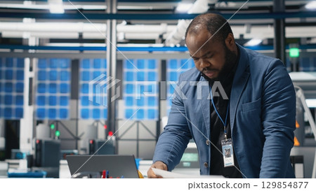 African american technician consulting performance metrics on papers and laptop, combining solar power expertise and efficiency in photovoltaics manufacturing industry. Camera B. African american technician consulting performance metrics on papers and laptop, combining solar power expertise and efficiency in photovoltaics manufacturing industry. Camera B. 129854877