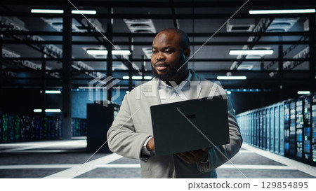 Engineer walking in server room backing up storage arrays using software on laptop. African american data center engineer scanning hardware logs for faults to fix bugs using device, camera A 129854895