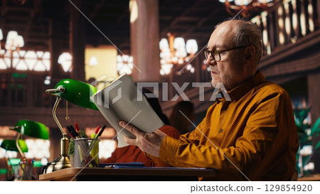 Elderly man immersed in reading and writing at a university campus library, working on his thesis with intellectual ambition and dedication to learning and academic achievement. 129854920