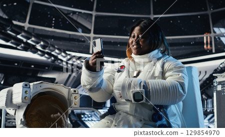 Cheerful female astronaut records selfie video with smartphone, speaking to subscribers during galactic mission. Jolly african american woman in starship recording content for followers Cheerful female astronaut records selfie video with smartphone, speaking to subscribers during galactic mission. Jolly african american woman in starship recording content for followers 129854970