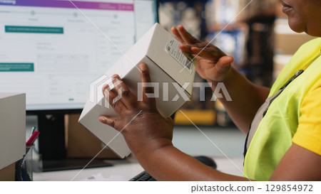 African american staff works on packages labeling for express delivery, ensuring accurate package tracking with a professional software. Worker processing orders in stock room. Camera B. 129854972