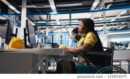 Photovoltaics factory worker with physical impairment using PC to monitor performance of solar panels manufacturing. Engineer works in disability friendly industrial facility. Camera B. 129854975