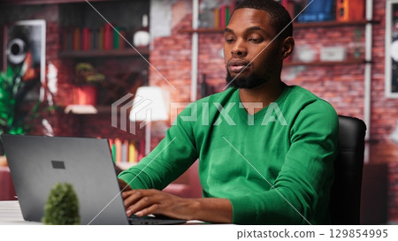 African american teleworker experiences lower back pain due to incorrect posture sitting at desk, dealing with office syndrome and muscle cramps. Young man feels overworked. Camera B. 129854995