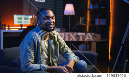 Portrait of black freelancer laying on the sofa and texting on phone, relaxing and smiling in his modern loft. Man solving daily teleworking tasks from the living room with cool lights. Camera A. 129855030