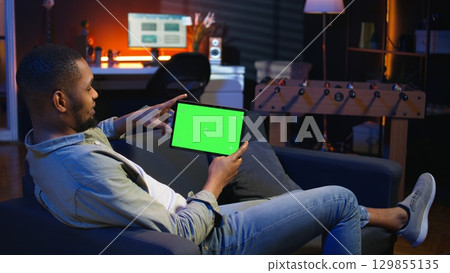 African american male freelancer holding phone with mockup copy space screen, relaxing on sofa in a space with neon lights and smart tv setup. Young person feels casual at home. Camera A. African american male freelancer holding phone with mockup copy space screen, relaxing on sofa in a space with neon lights and smart tv setup. Young person feels casual at home. Camera A. 129855135