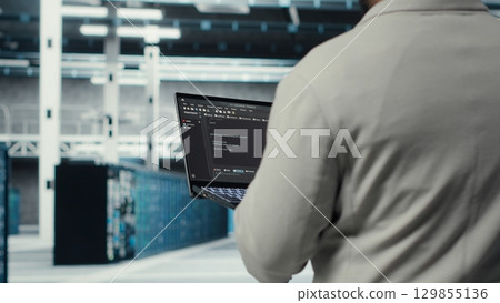 Engineer walking in data center using device to configure hardware optimized for AI tasks. Man doing maintenance on rigs performing load balancing for artificial intelligence computing, camera B 129855136