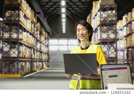 Asian worker blends retail and logistics operations with a laptop, processing custom orders and preparing parcels for shipment. Staff in hi-vis vest surrounded by cardboard boxes on racks. Asian worker blends retail and logistics operations with a laptop, processing custom orders and preparing parcels for shipment. Staff in hi-vis vest surrounded by cardboard boxes on racks. 129855182