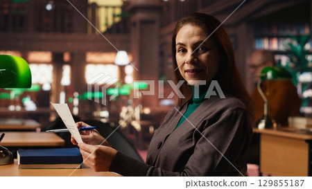 Woman with a serious expression studies papers at a public library desk, engaged in academic research and reading textbooks to enrich her understanding of literature. Self study. 129855187
