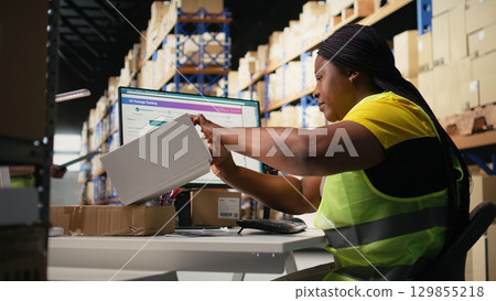 Black woman placing adhesive shipment tags on cardboard boxes, handles labeling and packaging tasks for order processing. Worker applying printed awb labels in storage depot. Camera A. 129855218