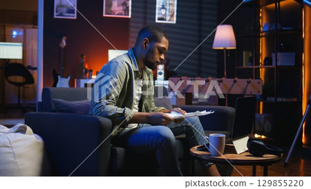 Young adult sitting on couch and reading research papers with analytics, multitasking with files and documents at home. Man diving into professional or personal topics of improvement. Camera A. Young adult sitting on couch and reading research papers with analytics, multitasking with files and documents at home. Man diving into professional or personal topics of improvement. Camera A. 129855220