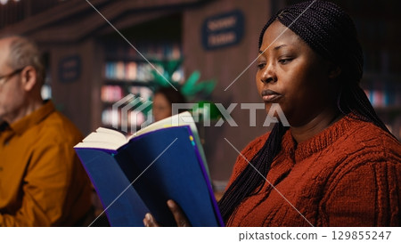 African american woman reading book and completing her class notes in a study room, preparing for her thesis and researching archived academic materials at the public library. African american woman reading book and completing her class notes in a study room, preparing for her thesis and researching archived academic materials at the public library. 129855247