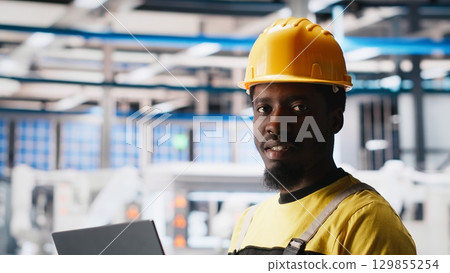Photovoltaics factory technician optimizing production flow using laptop, wearing hardhat. Sustainable energy solar panels plant specialist monitoring manufacturing process on device. Camera A. Photovoltaics factory technician optimizing production flow using laptop, wearing hardhat. Sustainable energy solar panels plant specialist monitoring manufacturing process on device. Camera A. 129855254