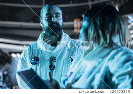 Team of astronauts verifying mission parameters on tablet to ensure safe operation during long distance space travel. Teamworking cosmonauts updating flight log during cosmic journey 129855259