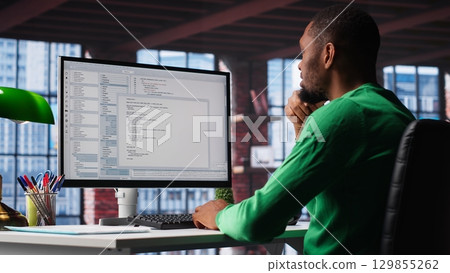 Black IT expert looking at user interface on computer at home office, typing and programming code on terminal window. Coding application used by technician writing script in loft. Camera B. 129855262