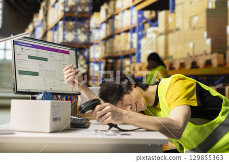 Tired employee deals with burnout and takes a nap with scanner in his hand, scanning shipping labels at warehouse desk. Order fulfillment and inventory management in a long shift. 129855363