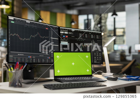 Green screen laptop in brokerage office next to workstation monitor displaying live stock charts. Mockup notebook on desk and computer with financial software in prop firm workspace 129855365