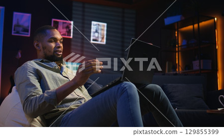 Black male freelancer taking online course to learn new skills from home, attend online video call and watching lecture from loft. Young man participates at a virtual training class. Camera A. Black male freelancer taking online course to learn new skills from home, attend online video call and watching lecture from loft. Young man participates at a virtual training class. Camera A. 129855399