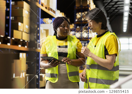 Diverse people processing e-commerce orders and reviewing invoices, organizing cargo goods inside industrial warehouse space filled with storage racks. Smooth delivery and quality control. Diverse people processing e-commerce orders and reviewing invoices, organizing cargo goods inside industrial warehouse space filled with storage racks. Smooth delivery and quality control. 129855415