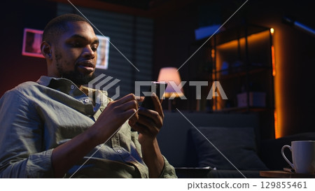 Young black guy texting his friends over social media messaging apps, using his smartphone to scroll on the internet. Focused person relaxing with his phone, sitting in his living room. Camera A. Young black guy texting his friends over social media messaging apps, using his smartphone to scroll on the internet. Focused person relaxing with his phone, sitting in his living room. Camera A. 129855461