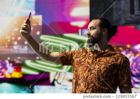Young relaxed man takes selfies and videos surrounded by fun neon lights and sound, representing the fusion of modern DJ culture, social media influence and digital art of nightlife performance. Young relaxed man takes selfies and videos surrounded by fun neon lights and sound, representing the fusion of modern DJ culture, social media influence and digital art of nightlife performance. 129855567
