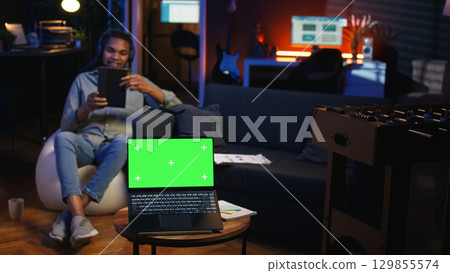 African american man browses phone next to mockup copy space while he lays on a bean bag, feeling happy and relaxed. Young teleworker in the living room feels pleased with remote job. Camera A. 129855574