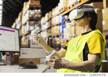 Asian industrial clerk uses VR glasses to streamline orders logistics in fulfillment center filled with big storage racks. Employee monitoring tracking software and packaging systems. Asian industrial clerk uses VR glasses to streamline orders logistics in fulfillment center filled with big storage racks. Employee monitoring tracking software and packaging systems. 129855579