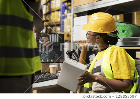 Black female worker chatting with CEO on web online conference, ensuring e-commerce retail activity on computer. Managing order fulfillment with awb tracking info in industrial warehouse. 129855597