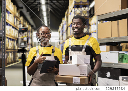 African American workers collaborating on a video marketing campaign in a storage room, fulfillment center retail operations. Coworkers live streaming vlog as commercial for large business. African American workers collaborating on a video marketing campaign in a storage room, fulfillment center retail operations. Coworkers live streaming vlog as commercial for large business. 129855684
