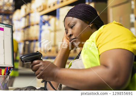 African american female napping on warehouse desk while she uses scanner, scanning shipping labels and barcode tags on a long shift. Tired overworked woman deals with burnout for e-commerce. 129855691