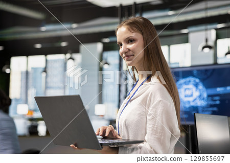 Smiling junior developer utilizing artificial intelligence to optimize performance using laptop. Cheerful young female engineer in office training machine learning models with notebook 129855697