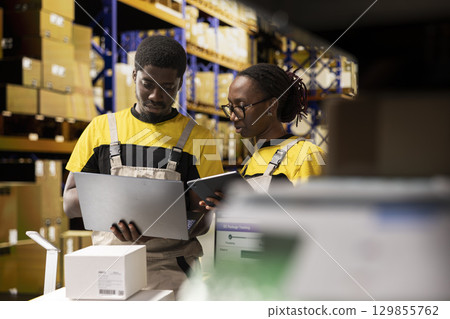 Warehouse workers overseeing parcels tracking info on laptop, preparing pallets for outbound shipment. Organized depot with storage racks, cardboard boxes and inventory processing tools. Warehouse workers overseeing parcels tracking info on laptop, preparing pallets for outbound shipment. Organized depot with storage racks, cardboard boxes and inventory processing tools. 129855762