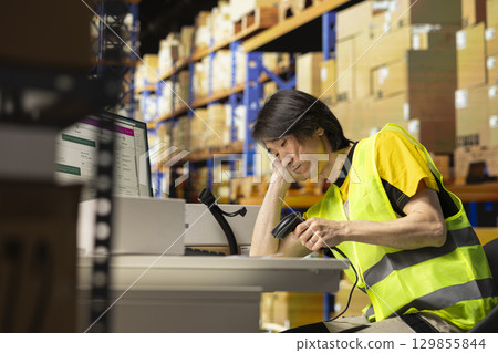 Sleepy asian staff member dealing with burnout in industrial warehouse, struggling to keep awake and ensure seamless package tracking service on software. Tired overworked employee. 129855844
